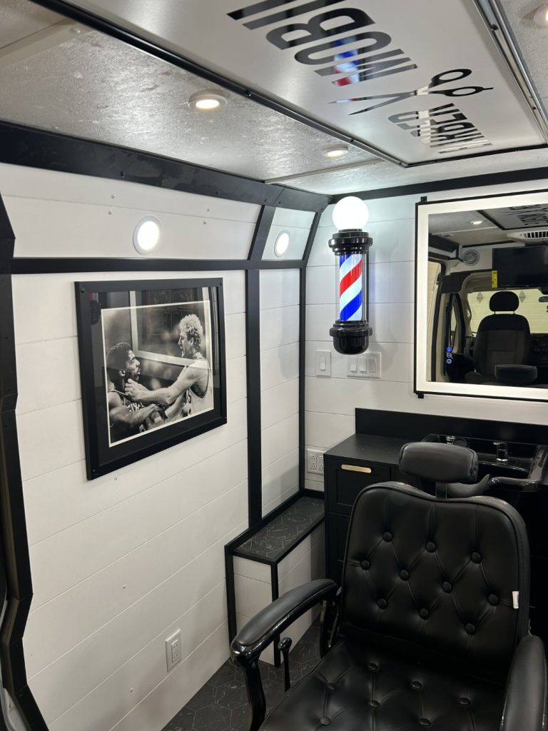 Inside the Barber Mobile with black leather barber chair, framed black and white sports photograph, illuminated barber pole, and backlit mirror built by Alexander's RV