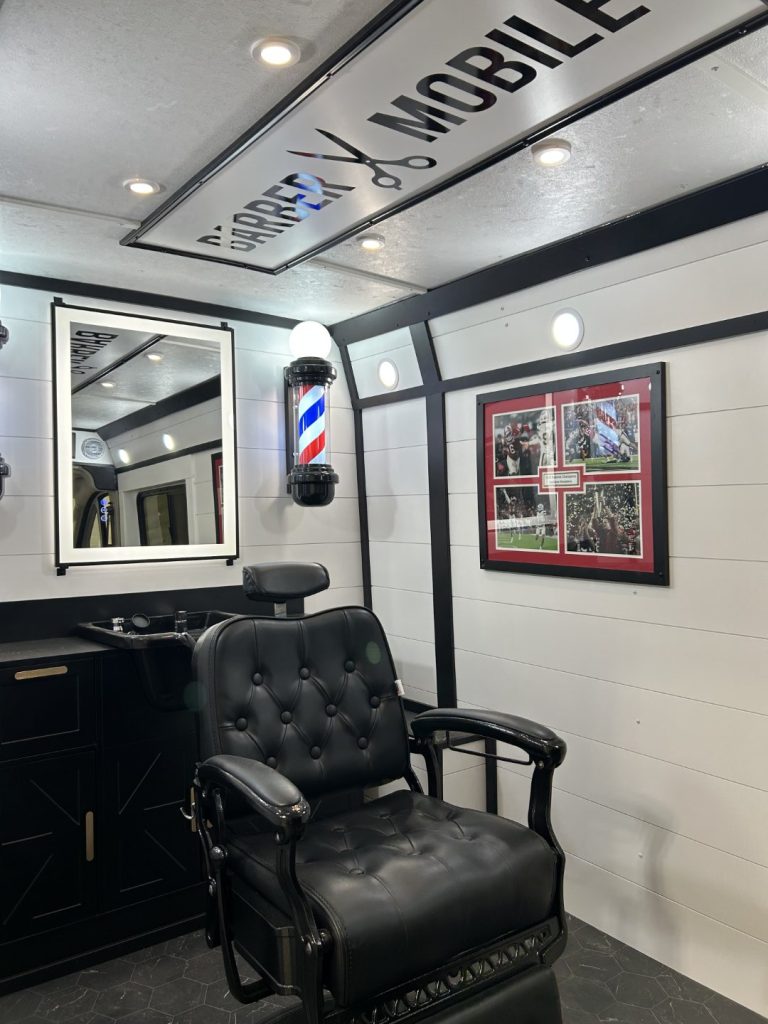 Inside the Barber Mobile with black leather barber chair, framed football memorabilia in red matting, barber pole, and backlit mirror built by Alexander's RV