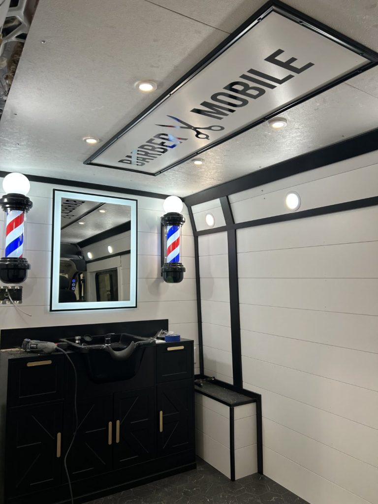 Black tufted leather barber chair inside the Barber Mobile with illuminated barber poles, backlit mirror, and shiplap walls built by Alexander's RV